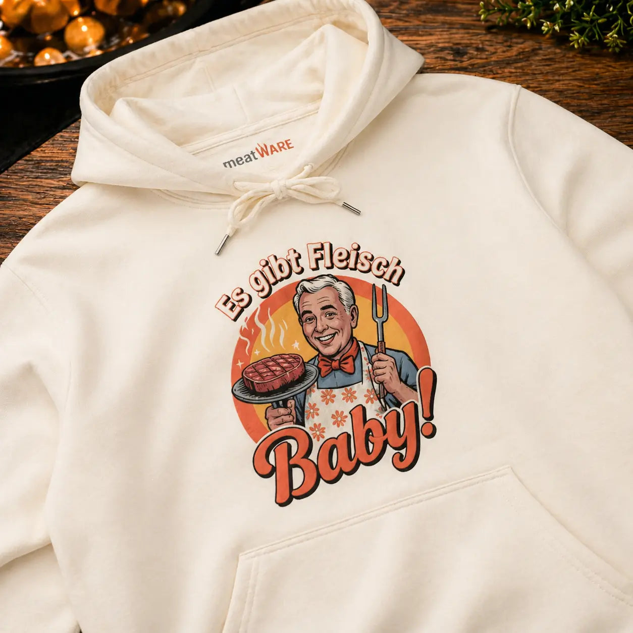 Premium Hoodie in Cream mit Retro Grill Design „Es gibt Fleisch Baby“ in Nahaufnahme auf Holztisch, Meatware Hoodie mit Steak Motiv und Grillmeister Illustration