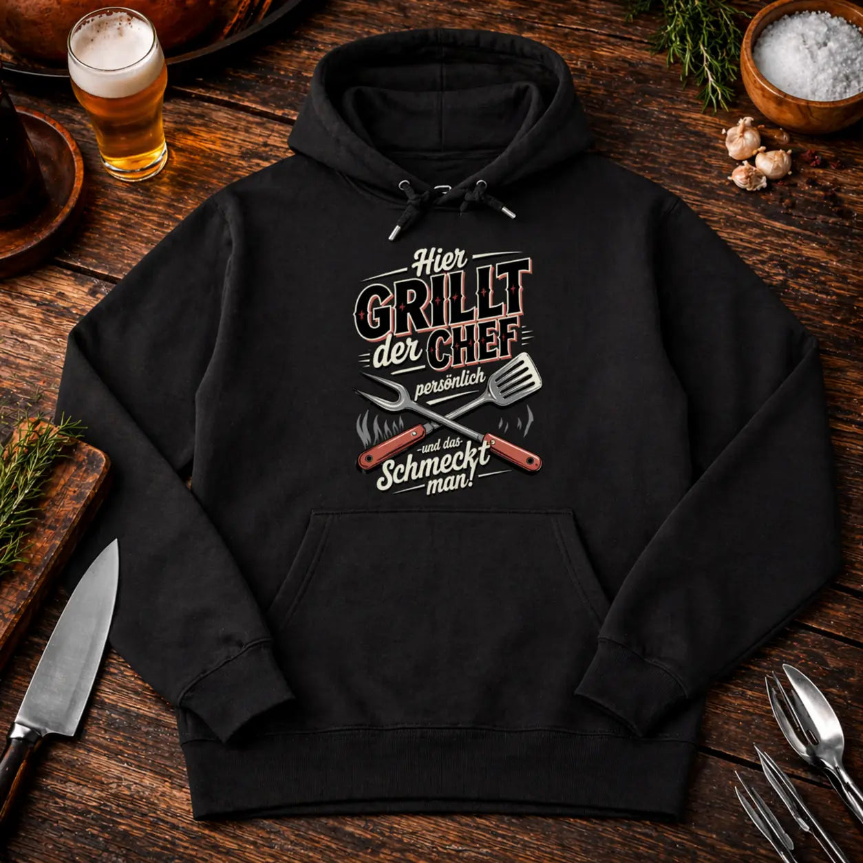 Schwarzer Premium Hoodie mit Aufdruck „Hier grillt der Chef persönlich und das schmeckt man“ auf rustikalem BBQ Holz Mockup mit Bier, Gewürzen und Grillbesteck