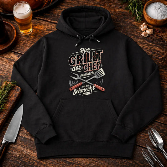 Schwarzer Premium Hoodie mit Aufdruck „Hier grillt der Chef persönlich und das schmeckt man“ auf rustikalem BBQ Holz Mockup mit Bier, Gewürzen und Grillbesteck
