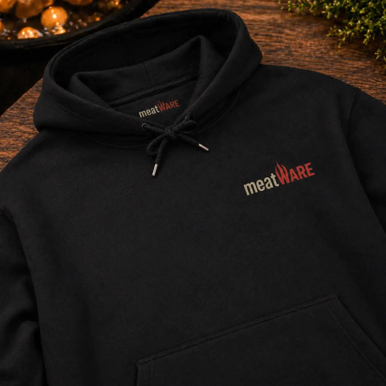 Schwarzer Premium Hoodie mit kleinem meatWARE Logo auf der Brust in Nahaufnahme auf Holztisch, minimalistischer Hoodie mit Kapuze, Kordeln und Nackenbranding