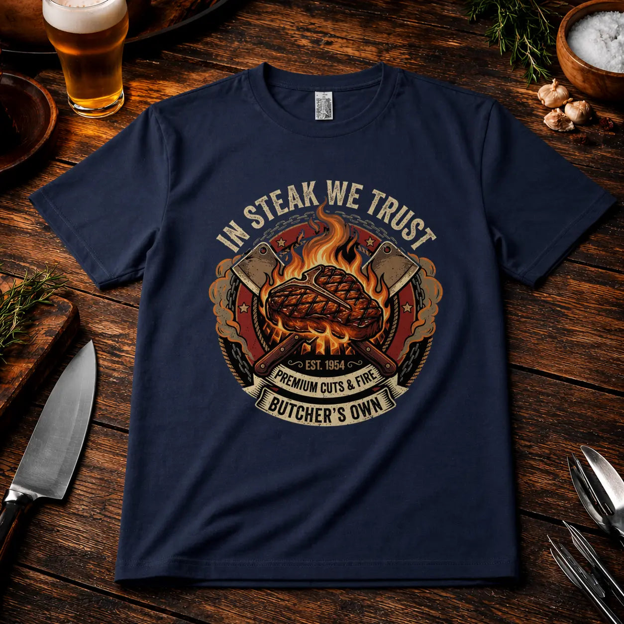 Navyfarbenes Premium T-Shirt mit „In Steak We Trust“ Grillmotiv im rustikalen BBQ Mockup auf Holztisch mit Bier, Messer und Gewürzen, realistische Stoffstruktur und hochwertiger Druck.