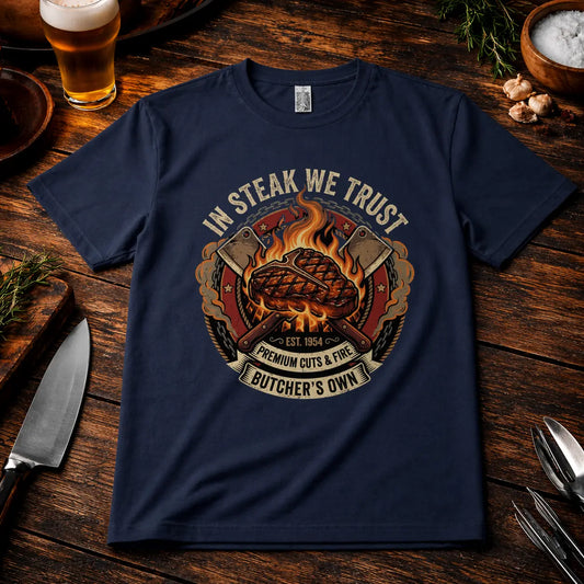 Navyfarbenes Premium T-Shirt mit „In Steak We Trust“ Grillmotiv im rustikalen BBQ Mockup auf Holztisch mit Bier, Messer und Gewürzen, realistische Stoffstruktur und hochwertiger Druck.