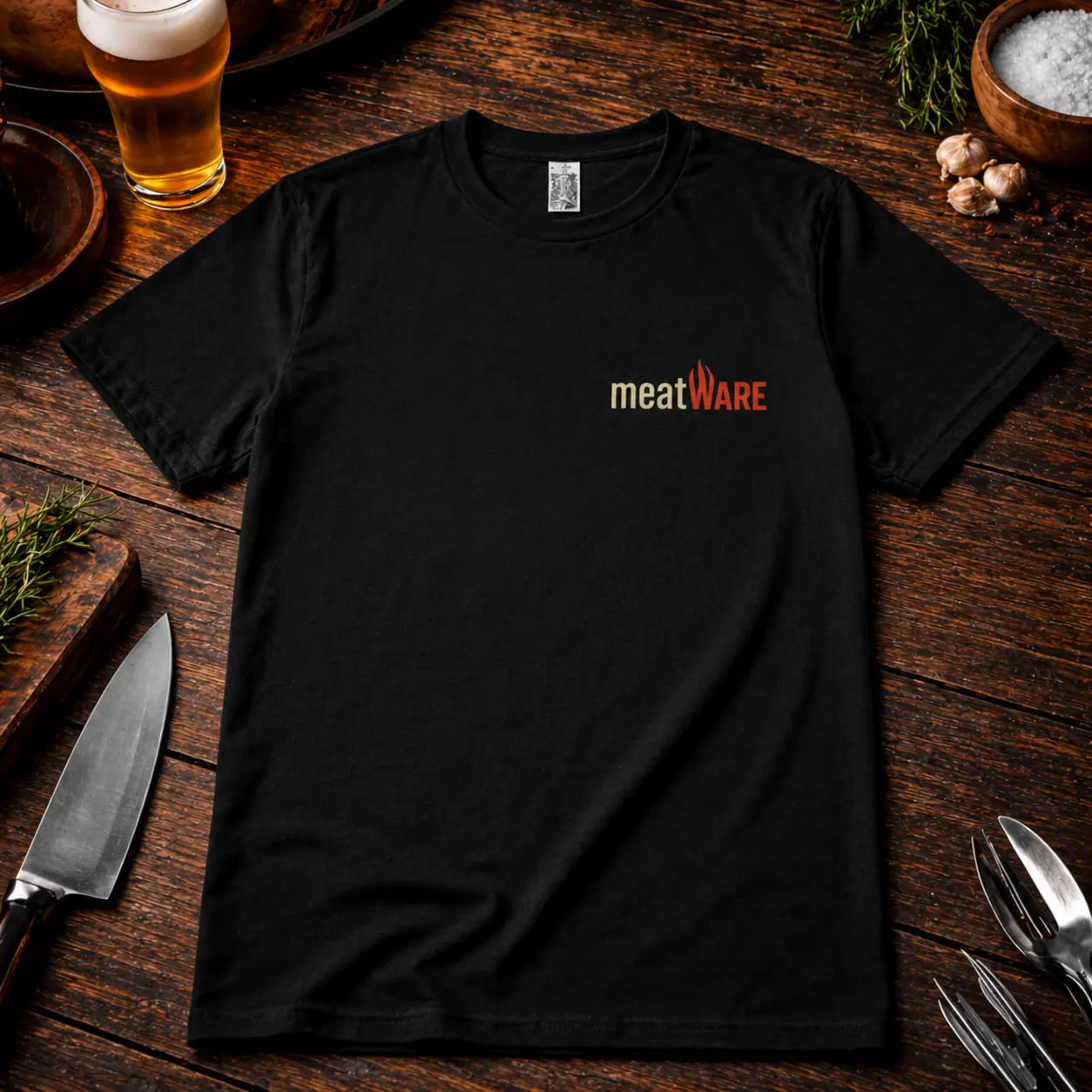 Schwarzes Premium T-Shirt mit kleinem Meatware Logo auf der Brust im rustikalen BBQ Mockup auf Holztisch mit Bier, Messer und Kräutern, realistische Stoffstruktur und hochwertiger Druck.
