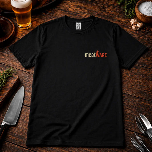 Schwarzes Premium T-Shirt mit kleinem Meatware Logo auf der Brust im rustikalen BBQ Mockup auf Holztisch mit Bier, Messer und Kräutern, realistische Stoffstruktur und hochwertiger Druck.