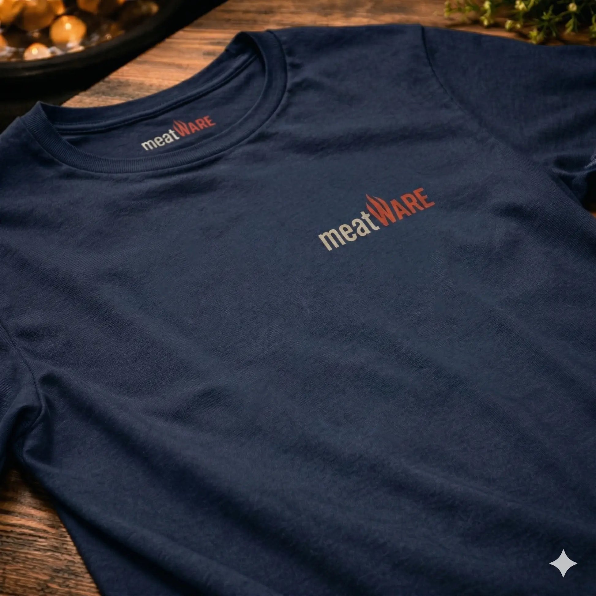 Nahaufnahme eines dunkelblauen T-Shirts, das flach auf einem rustikalen Holztisch liegt. Auf der linken Brustseite ist das "meatWARE"-Logo mit einer kleinen Flamme in Beige und Rot aufgedruckt. Das gleiche Logo ist auch als Etikett im Kragen sichtbar. Der Hintergrund ist leicht unscharf mit warmen Lichtern und etwas Grünpflanze, passend zu einem Grill- oder Küchenthema.