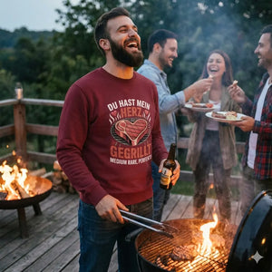 Mann in weinrotem Organic Sweatshirt mit Steak-Herz-Motiv "Du hast mein Herz gegrillt" genießt BBQ-Abendstimmung.