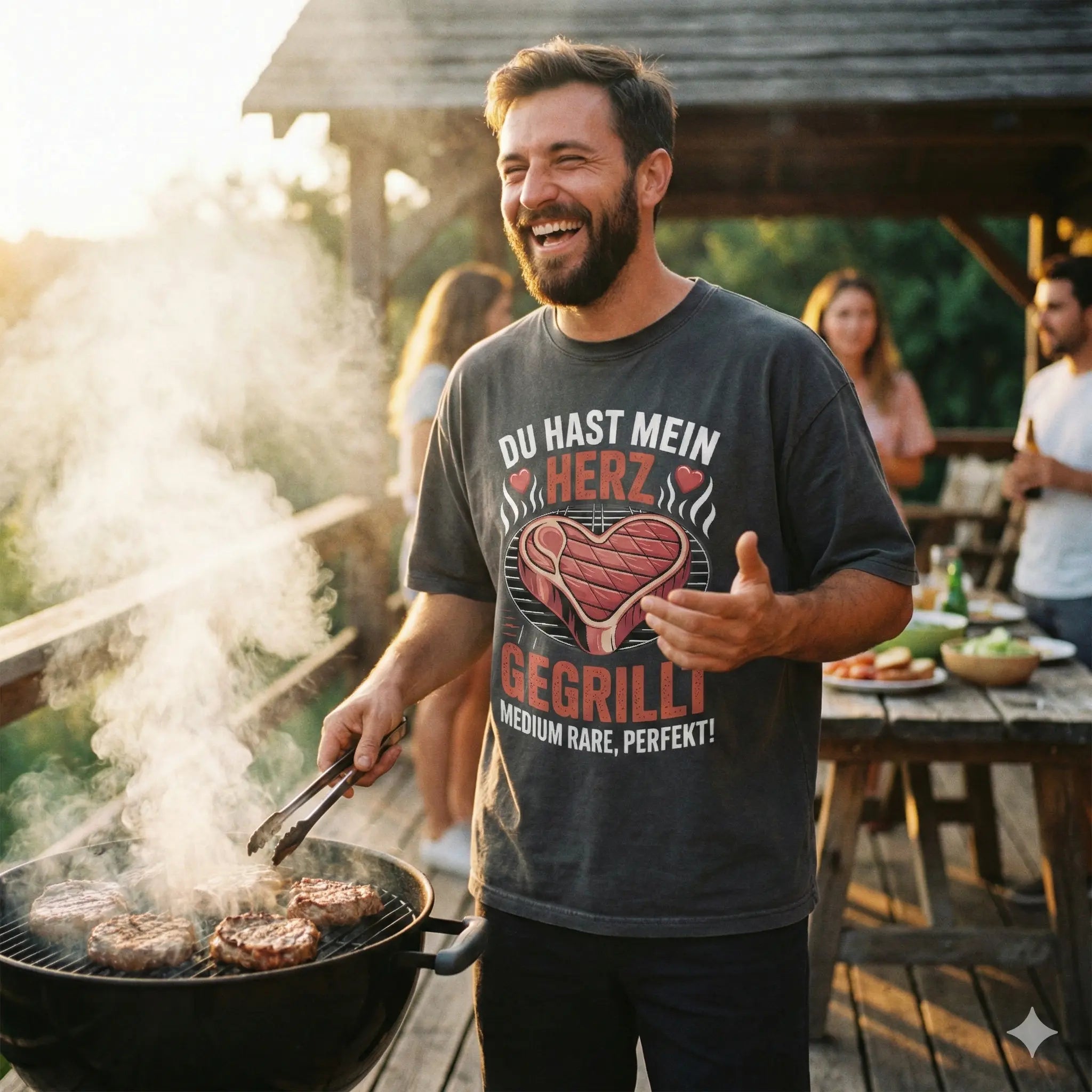 Mann in dunkelgrauem Acid-Wash Oversized Shirt mit Steak-Herz-Motiv steht lachend am Grill, warme Abendstimmung