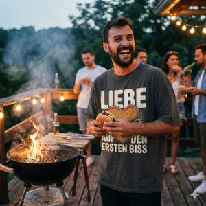 Lachender Mann auf Grillparty trägt graues Oversized Shirt mit Burger-Herz-Motiv "Liebe auf den ersten Biss", warme Abendstimmung