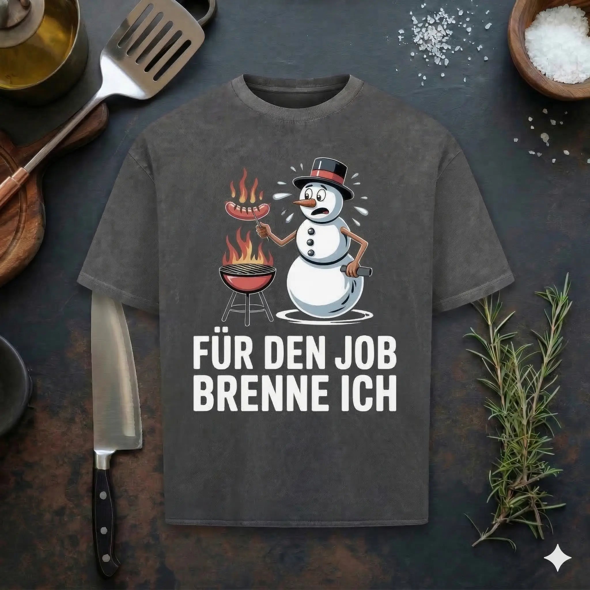 Ein flach liegendes, dunkelgraues T-Shirt mit Stone-Wash-Optik auf einem rustikalen dunklen Holztisch, umgeben von einem Kochmesser, frischem Rosmarin, einer Holzschale mit Meersalzflocken, einem Mörser mit Stößel und einem Pfannenwender. Auf dem T-Shirt befindet sich ein Cartoon-Druck eines Schneemanns mit Zylinder, der schwitzt und Wasser verliert, während er eine Wurst über einem brennenden Kugelgrill brät. Darunter steht der deutsche Text in weißer Schrift: „FÜR DEN JOB BRENNE ICH“.
