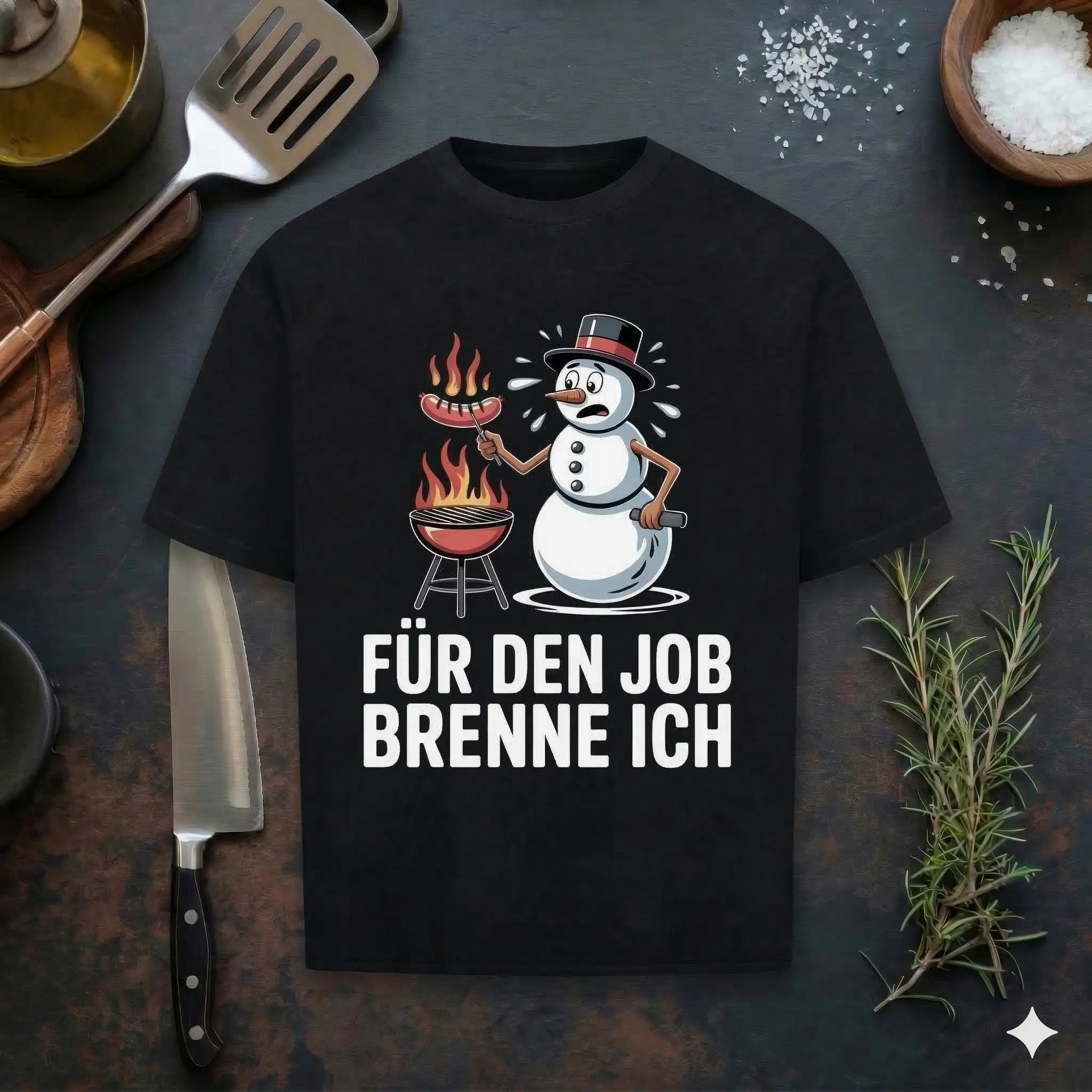 Ein schwarzes T-Shirt in einem Flachbett-Mockup, bedruckt mit einem Cartoon-Schneemann in einem Zylinder, der eine Wurst über einem brennenden Kugelgrill brät. Darunter steht der weiße Text „FÜR DEN JOB BRENNE ICH“. Das Shirt liegt auf einer dunklen Schieferplatte, umgeben von einem Kochmesser, einem Pfannenwender, frischem Rosmarin, Meersalz und Öl.