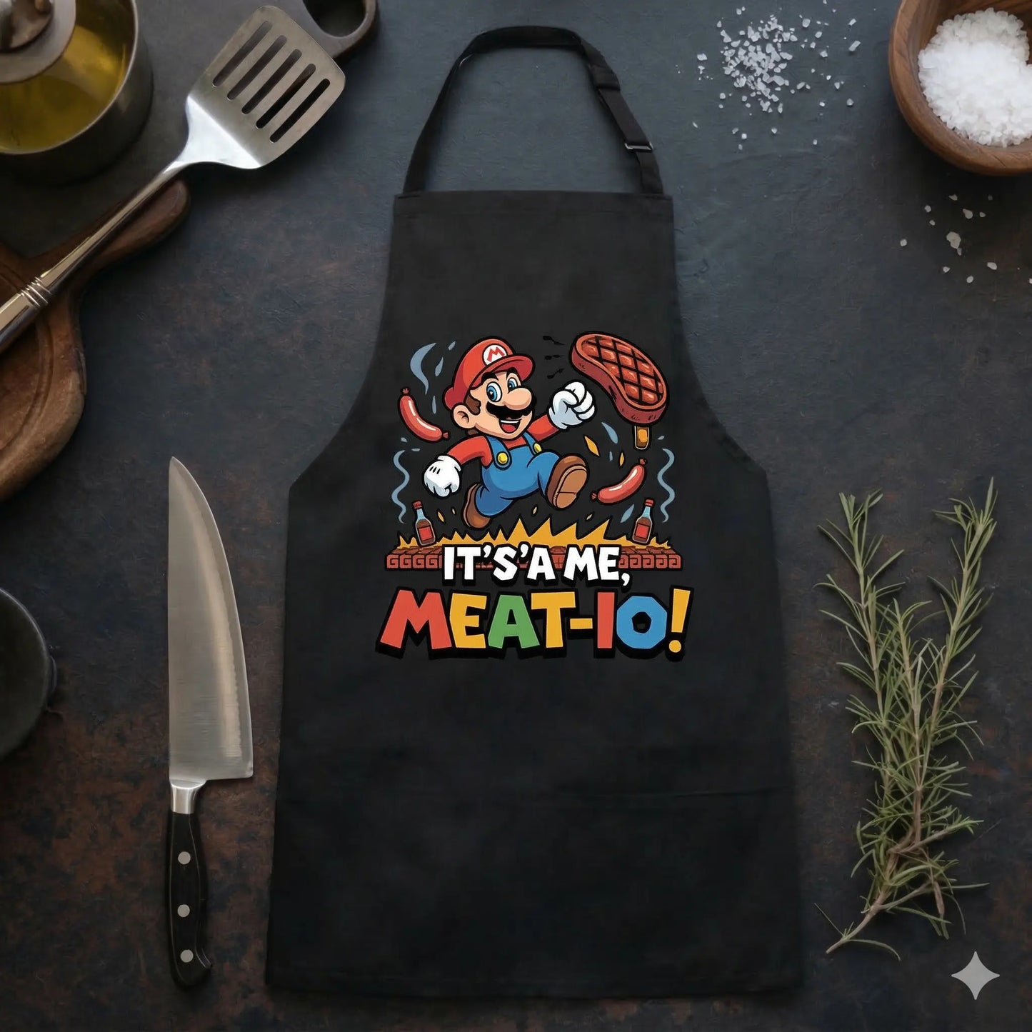 Ein Foto aus der Vogelperspektive, das eine schwarze Grillschürze zeigt, die flach auf einer dunklen Küchenarbeitsplatte liegt. Die Schürze ist mit einem farbenfrohen Aufdruck versehen, der eine Karikatur von Super Mario zeigt, der ein großes Stück Fleisch über einem brennenden Grill hält, umgeben von Würstchen. Darunter steht der Text: "IT'S'A ME, MEAT-IO!". Die Schürze ist umgeben von verschiedenen Küchenutensilien: links liegen Holzschneidebretter, ein großes Kochmesser, ein Pfannenwender aus Metall und 