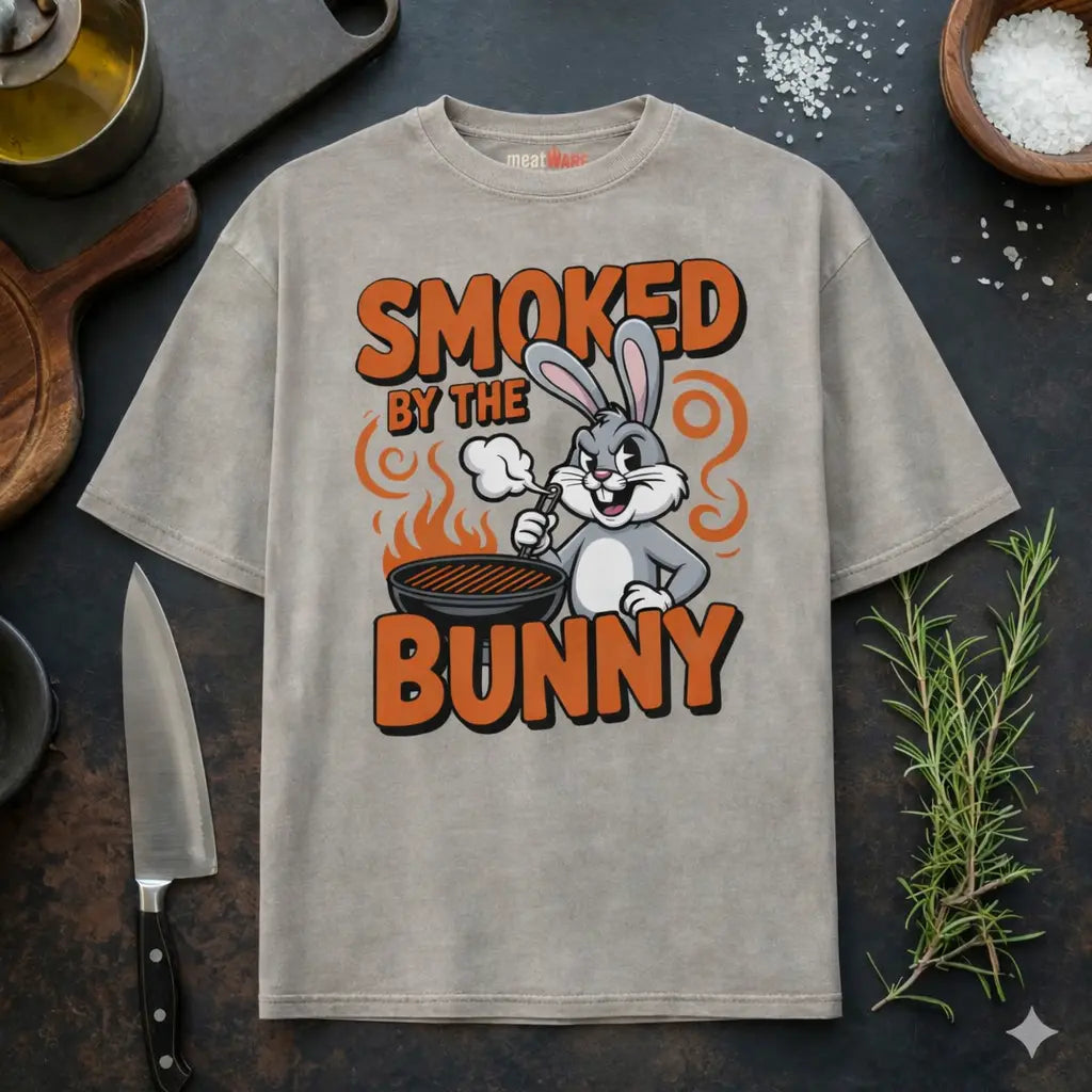Beiges Meatware Oversized T-Shirt mit Smoked by the Bunny BBQ Hasen Grill Design und Meatware Nackenbranding auf dunklem Küchen Hintergrund mit Messer, Rosmarin und grobem Salz