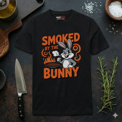Schwarzes Meatware Premium T-Shirt mit Smoked by the Bunny BBQ Hasen Grill Design und Meatware Nackenbranding auf dunklem Küchen Hintergrund mit Messer, Rosmarin und Gewürzen