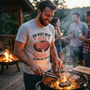 Lachender Mann am Grill in grauem T-Shirt mit Herz-Steak-Motiv "Du hast mein Herz gegrillt".