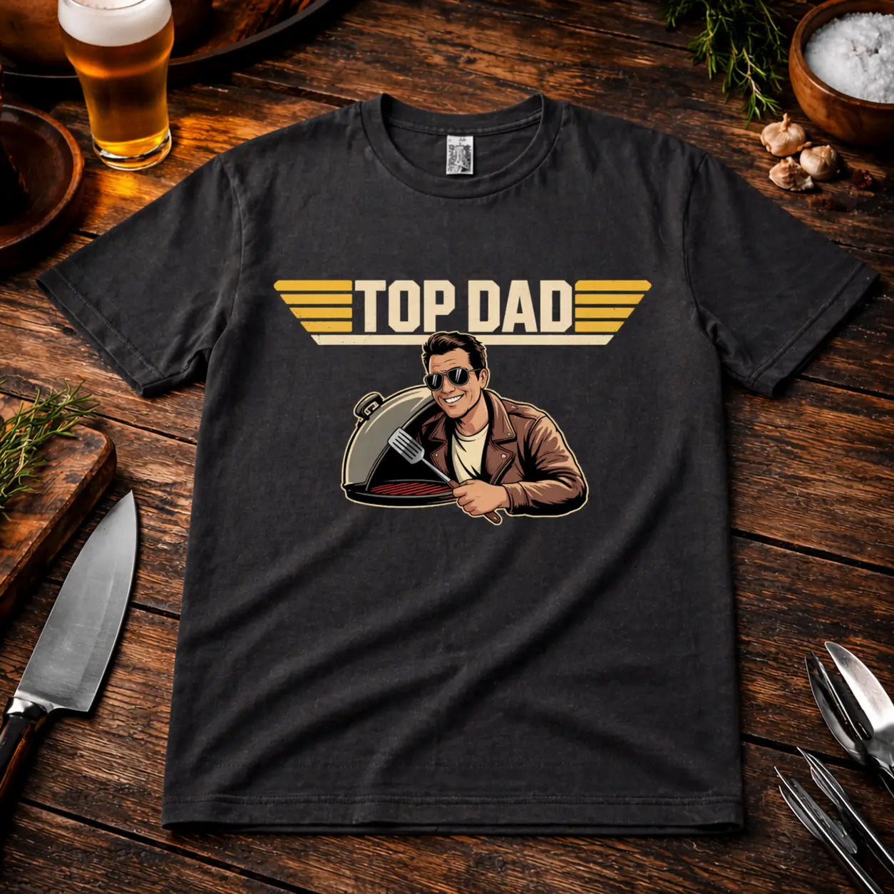 Schwarzes Premium T-Shirt mit „Top Dad“ Grillmotiv im realistischen BBQ Mockup auf rustikalem Holztisch mit Bier, Kräutern, Gewürzen und Grillzubehör – ideales Vatertagsgeschenk für Grillfans