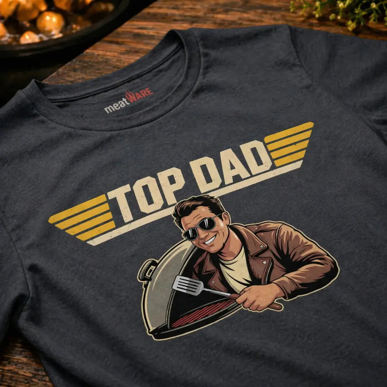 Schräg fotografierte Nahaufnahme eines washed schwarzen Premium T-Shirts mit „Top Dad“ Grillmotiv und sichtbarem Meatware Nackenbranding im rustikalen BBQ Mockup auf Holzoberfläche als Vatertags Geschenkidee